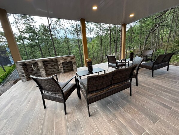 Un patio al aire libre con chimenea de piedra, mobiliario de mimbre y vista a un bosque.