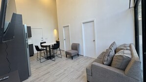 Un salon moderne avec un canapé gris, une petite table entourée de chaises et un téléviseur posé sur un meuble.