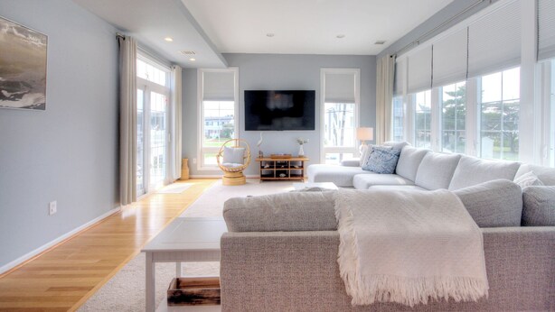 A spacious living room with a grey sofa, a white coffee table, and a flat-screen TV mounted on the wall. - Bay Avenue Bliss, Newly Updated, Spacious Yard, OCNJ South Shore (Ocean City)