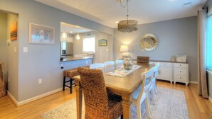 A dining area with a wooden table, wicker chairs, a chandelier, and a kitchen in the background. - Bay Avenue Bliss, Newly Updated, Spacious Yard, OCNJ South Shore (Ocean City)