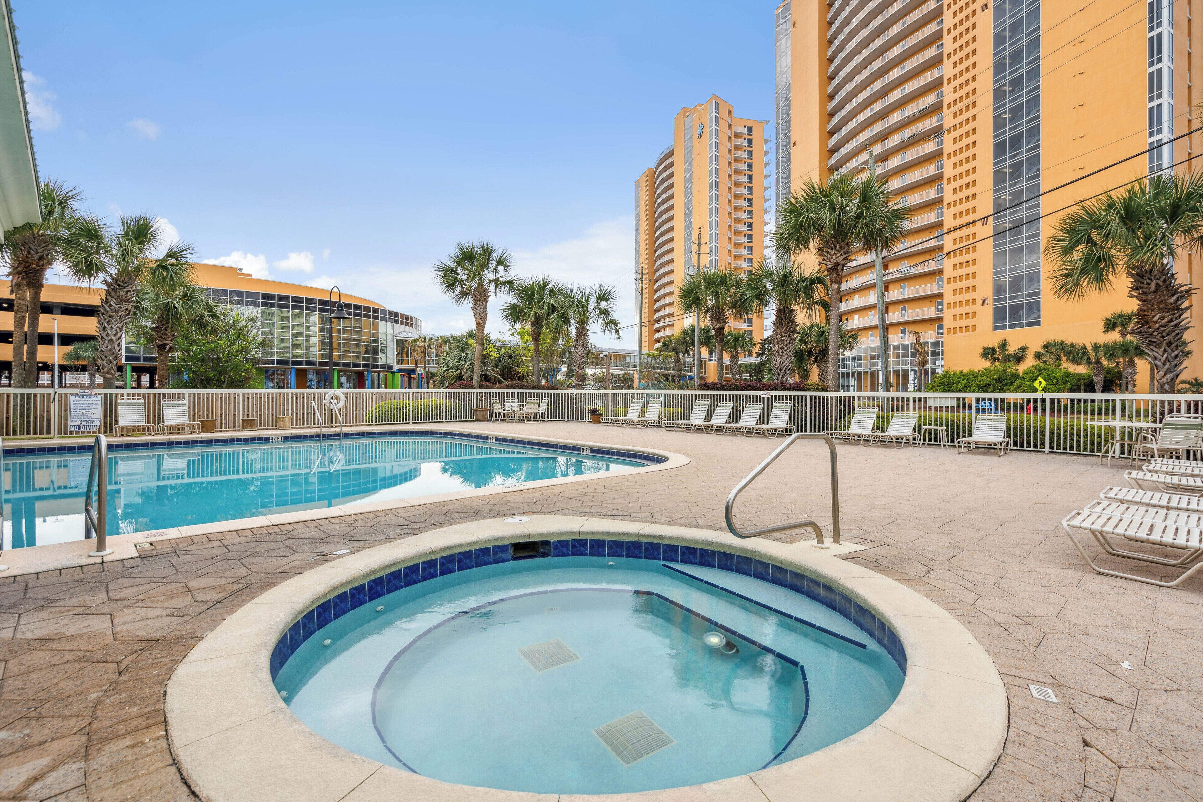 Un espace piscine d’hôtel comprenant un jacuzzi, des chaises longues et une allée bordée de palmiers.