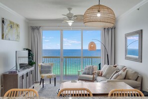 A balcony with a view of the ocean, a ceiling fan, a woven light fixture, a sofa, a chair, a dining table, and a TV on a cabinet.
