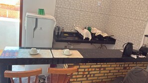 Eine Küchenzeile mit Kühlschrank, Spüle und einer Arbeitsplatte, auf der ein Kaffeeautomat und ein Wasserkocher stehen. Im Vordergrund befinden sich Stühle und ein Tisch.