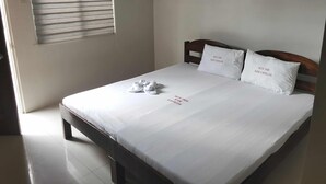 Una cama bien hecha con sábanas blancas y dos almohadas, cabecera de madera, ventana con persianas y un equipo de aire acondicionado en la pared.
