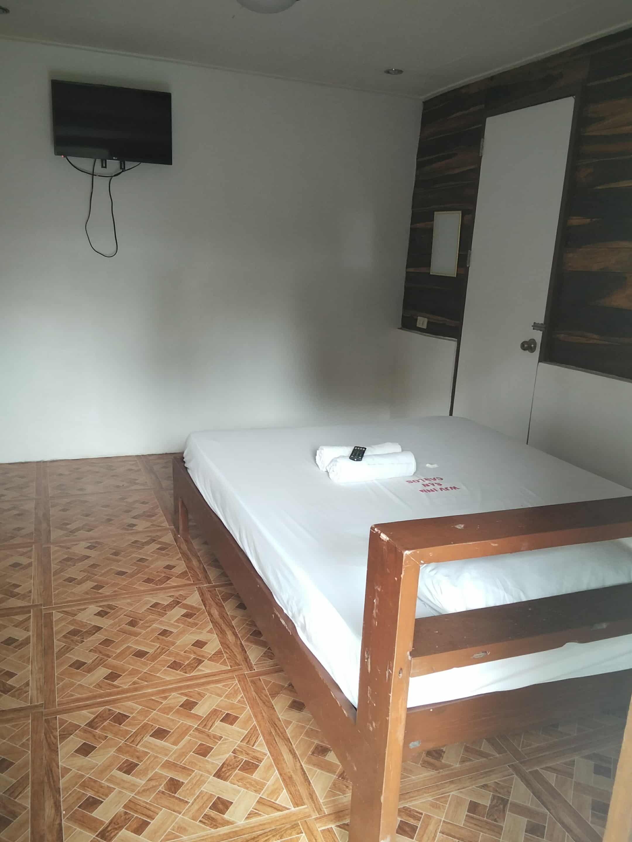 Una cama individual con estructura de madera, colchón blanco y una toalla doblada con un control remoto encima. Hay un televisor instalado en la pared y una puerta con diseño de panel de madera.