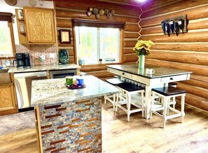 Eine rustikale Kücheneinrichtung im Blockhausstil mit einer Steininsele, Holzschränken und einem Essbereich mit weißen Stühlen und einem Tisch.