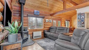 A log cabin interior with a fireplace, leather sofa, and a large window. - Log Cabin: Hot Tub, Sauna, Wet Bar & Mtn Views (Missoula County)