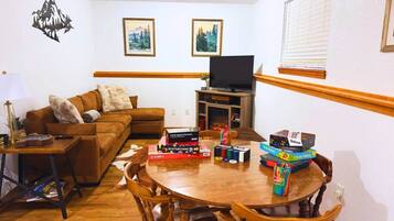 Una sala de estar acogedora con un sofá marrón, una mesa de centro de madera y un televisor sobre un mueble. Hay cuadros en la pared y una ventana con persianas.