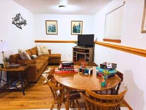 Ein gemütliches Wohnzimmer mit braunem Sofa, einem Holztisch und einem Fernseher auf einem Schrank. An der Wand hängen Bilderrahmen, und es gibt ein Fenster mit Jalousien.
