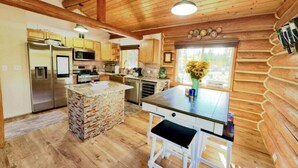 A rustic cabin-style kitchen with wooden walls, a stone island, and a large window with a view. - Log Cabin: Hot Tub, Sauna, Wet Bar & Mtn Views (Missoula County)