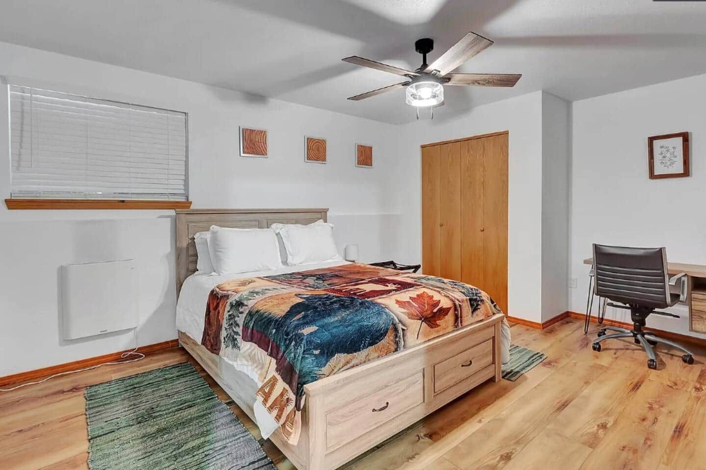 Quarto com cama de madeira, escrivaninha, cadeira, ventilador de teto e janela com persianas.