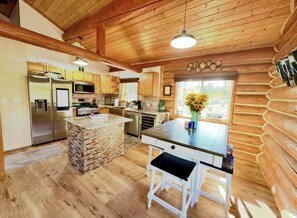A rustic cabin-style kitchen with wooden walls, a stone island, and a large window with a view. - Log Cabin: Hot Tub, Sauna, Wet Bar & Mtn Views (Missoula County)