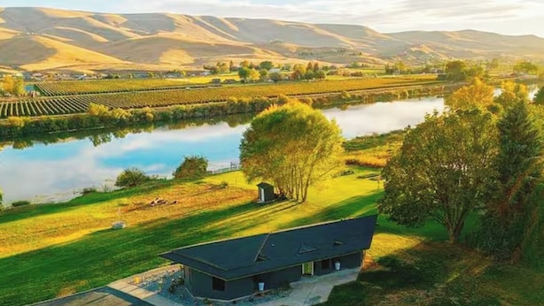 Uma casa moderna com telhado plano, localizada perto de um corpo d'água e rodeada por uma paisagem gramada com colinas ao fundo.
