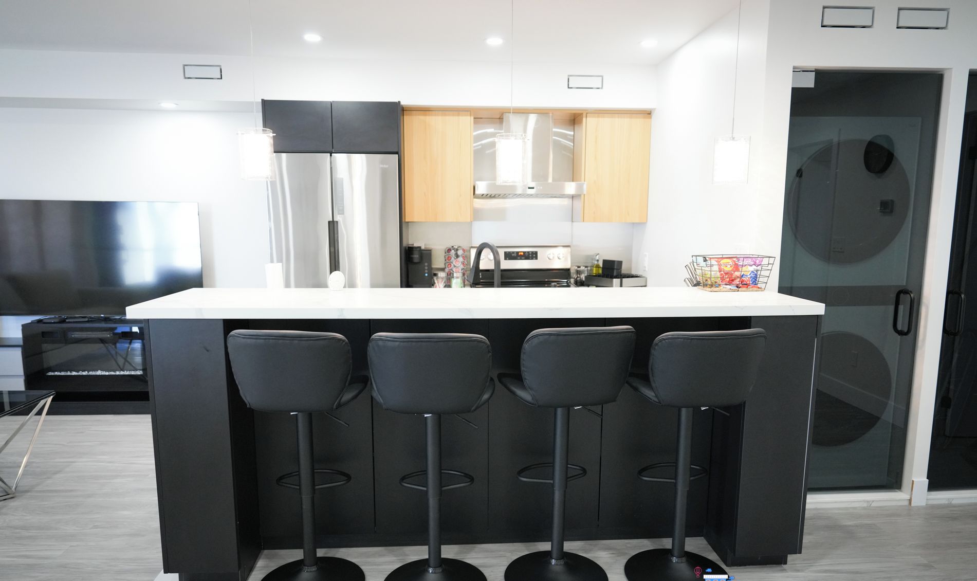 A modern kitchen with a central island, four bar stools, stainless steel appliances, and wooden cabinets.