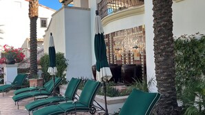 A courtyard with lounge chairs, palm trees, and a building with a tiled roof and balcony.