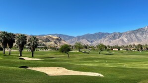 A golf course with sand bunkers, palm trees, and mountains in the background.