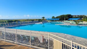 A large outdoor swimming pool with multiple lanes, surrounded by lounge chairs and umbrellas, and a wooden deck in the foreground.