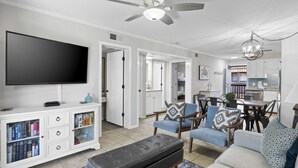 A modern living room with a flat-screen TV, a white entertainment unit with books, a grey bench, and a dining area with chairs and a table.