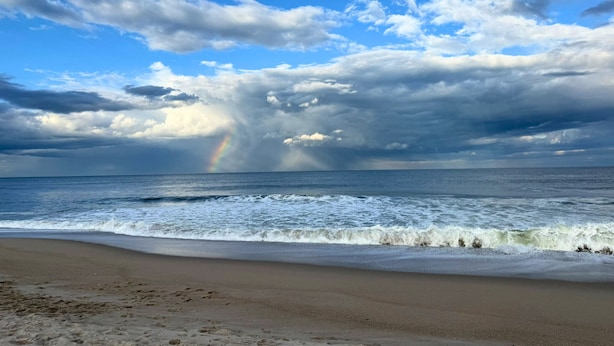 A beach with waves, a rainbow, and a cloudy sky.