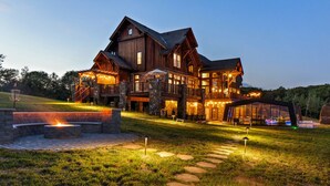Una casa grande de madera de varios pisos con base de piedra, luces exteriores y una fogata.