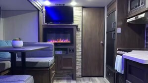 A compact kitchen with a built-in TV, a wood stove, and a dining area with a sofa. - The GreyWolf - Convenient & Cozy RV (Mobile)