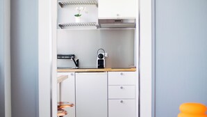 Kapea käytävä johtaa pieneen keittiöön, jossa on valkoiset kaapit, puinen työtaso ja pieni jakkara.