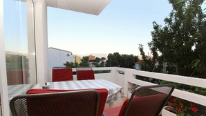 A balcony with a table and chairs, a potted plant with red flowers, and a view of buildings and trees. - Holiday apartment with air conditioning (Baška)
