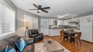 A modern living room with a leather sofa, a coffee table, and a dining area with chairs. A ceiling fan and a kitchen with white cabinets and appliances are visible. - Knockan Hill Rentals - Ground Floor Suite (Victoria)