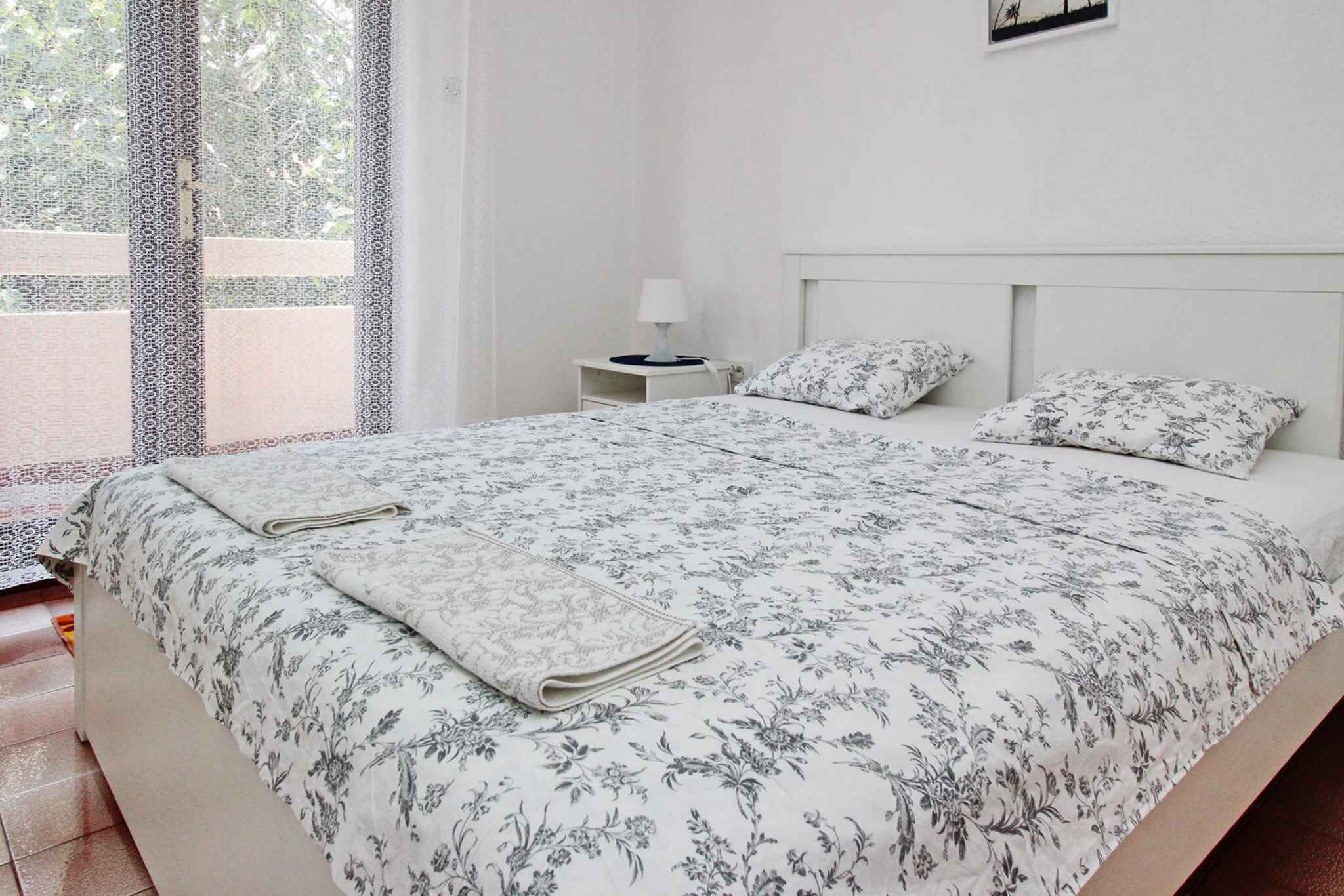 A neatly made bed with a floral bedspread and matching pillows, a bedside table with a lamp, and a window with a view of greenery.