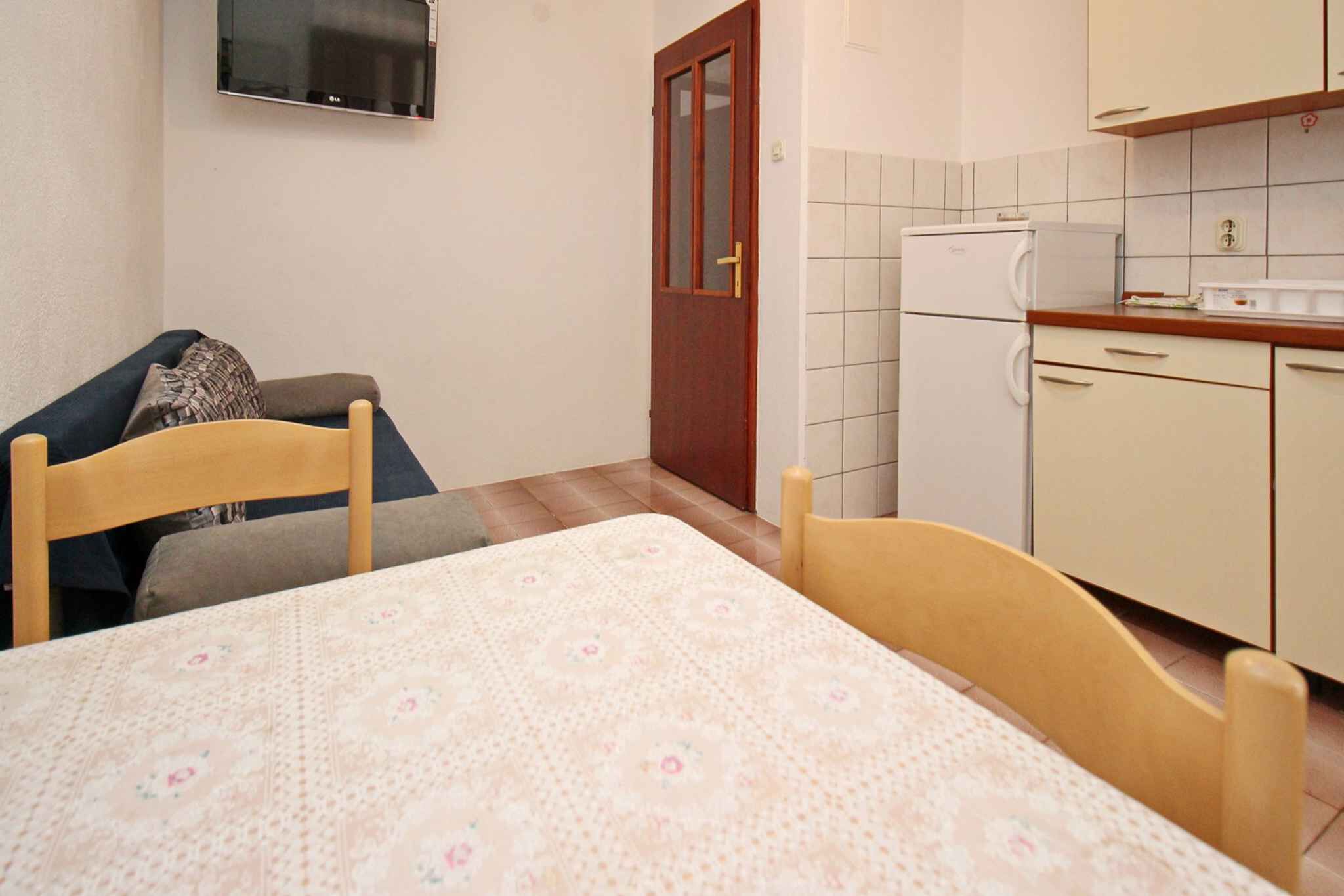 Een kleine, compacte kamer met een bed, een tafel, een stoel, een bank, een televisie en een kitchenette met een koelkast en kastjes.