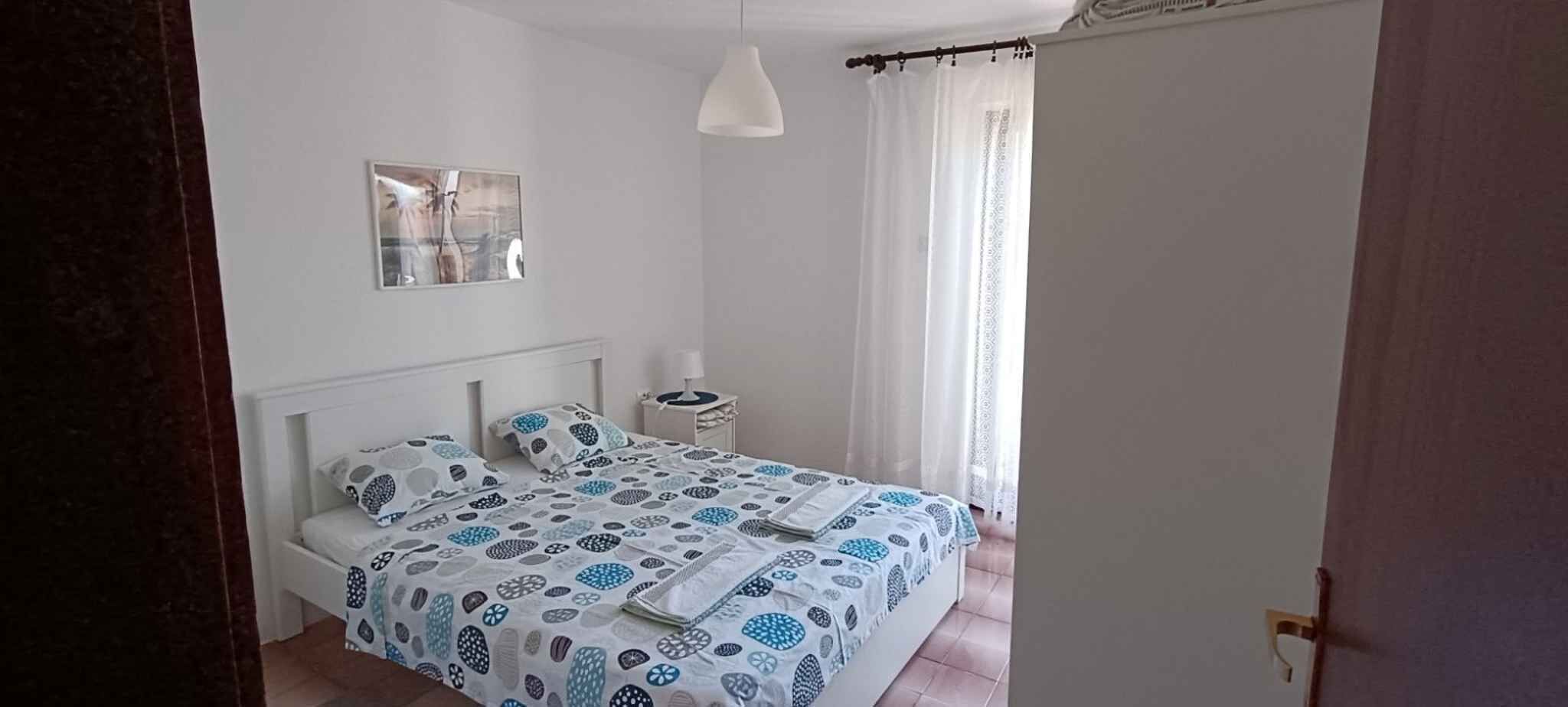 Un dormitorio con una cama, una mesita de noche, una lámpara y una ventana con cortinas.