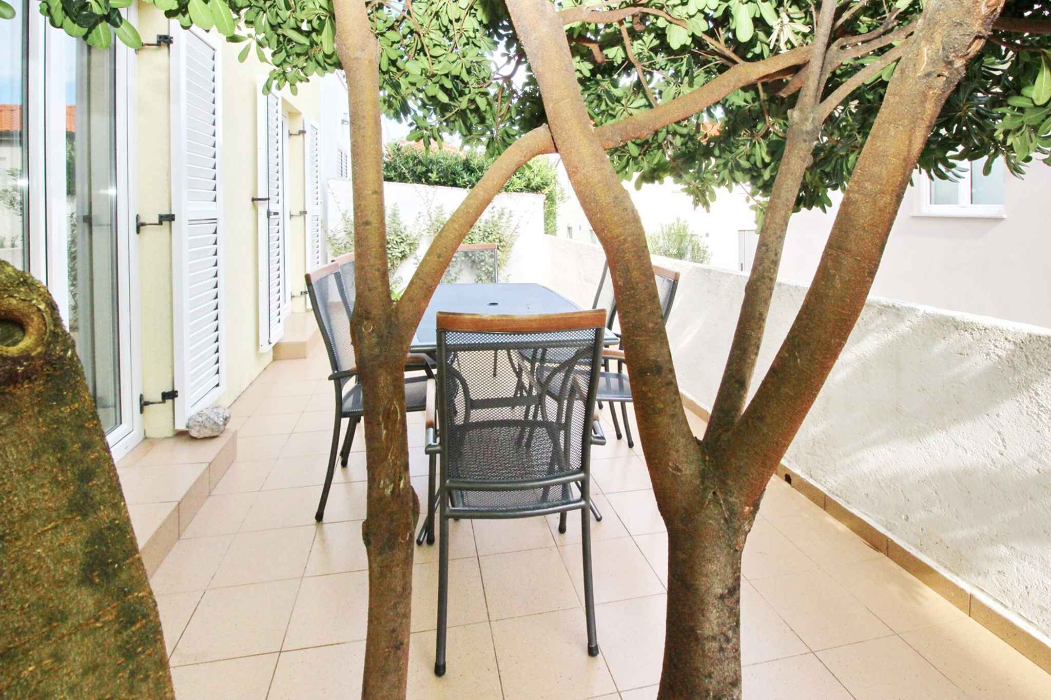 Eine Terrasse im Freien mit Tisch und Stühlen, umgeben von Bäumen und einer weißen Mauer.