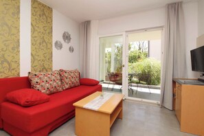 Un canapé rouge agrémenté de coussins à motifs, une table basse en bois, un téléviseur et une porte-fenêtre coulissante donnant sur un espace extérieur avec une table et des chaises.