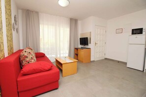 A living room with a red sofa, a wooden coffee table, a television, and a kitchen area with a microwave and refrigerator.