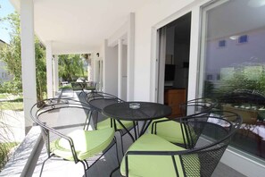Un espace extérieur aménagé, abrité et doté de tables et de chaises, une porte-fenêtre coulissante donnant sur une cuisine et une vue sur la verdure environnante.