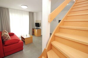 A modern living room with a red sofa, a wooden coffee table, and a TV stand. There is a wooden staircase leading to another level.