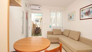 Ein Wohnzimmer mit einem runden Holztisch, zwei Stühlen, einem beigefarbenen Sofa und einem Fenster mit weißen Vorhängen.