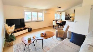 Ein modernes Wohnzimmer mit Flachbildfernseher, einem Essbereich mit Tisch und Stühlen sowie einer Küche im Hintergrund.