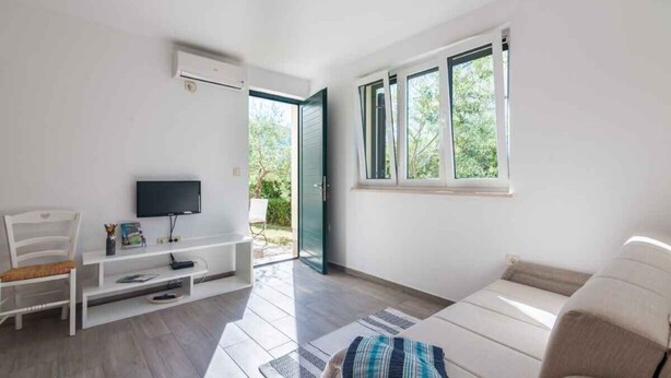 A modern living room with a flat-screen TV, white furniture, a sofa, and large windows offering a view of greenery. - Holiday apartment with shared-use pool (Jurandvor)