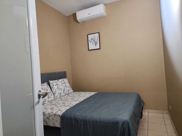 Et lille værelse med en enkelt seng, gråt sengegavl, et billede i ramme på væggen og en aircondition-enhed over sengen.