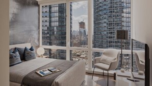 Ein modernes Schlafzimmer mit einem großen Bett, einem Sessel und Blick auf eine Skyline durch große Fenster.