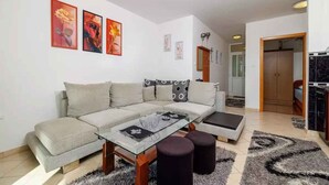 Una sala de estar moderna con un sofá, una mesa de centro y otomanas. Hay cuadros enmarcados en la pared y una zona de cocina al fondo.