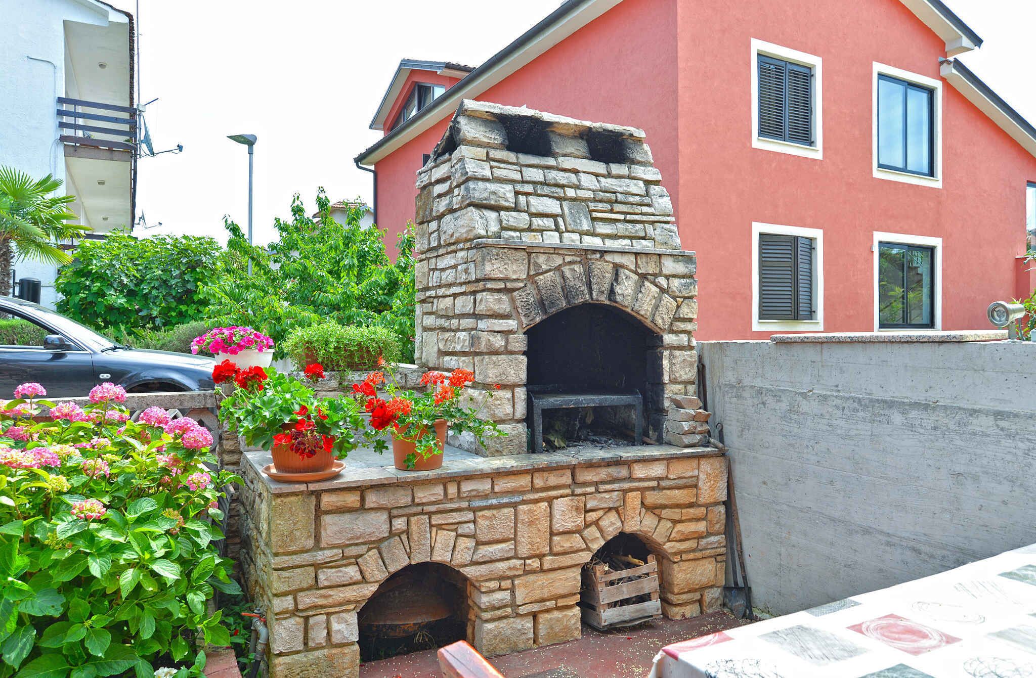 A stone-built outdoor kitchen with a chimney, surrounded by potted plants and a pink building in the background.
