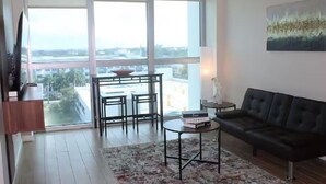 A modern living room with a large window, a black leather sofa, a small round table, and a TV on the left. - The Oasis Downtown Fort Myers/Pool View Retreat (Fort Myers)