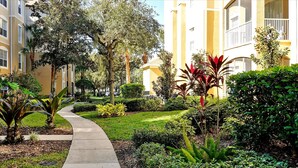 Un sentier traverse un jardin bien entretenu, où l’on trouve une variété de plantes et d’arbres, et il est bordé de bâtiments dotés de balcons.