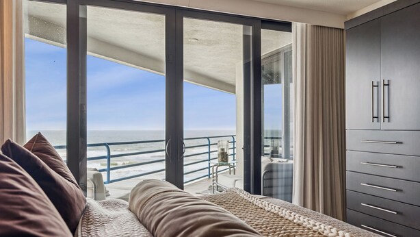 A balcony with a view of the sea, a sofa with cushions, a glass door, and a wardrobe with drawers. - Ebb Tide - 703 your oceanfront dream vacation destination awaits! (New Smyrna Beach)