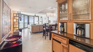 A modern kitchen with wooden cabinets, a dishwasher, and a coffee maker. There is a dining area with a table and chairs, and a large window offering a view. - Ebb Tide - 703 your oceanfront dream vacation destination awaits! (New Smyrna Beach)