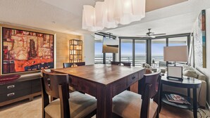 A modern dining area with a wooden table and chairs, a large abstract painting on the wall, and a view of the ocean through large windows. - Ebb Tide - 703 your oceanfront dream vacation destination awaits! (New Smyrna Beach)