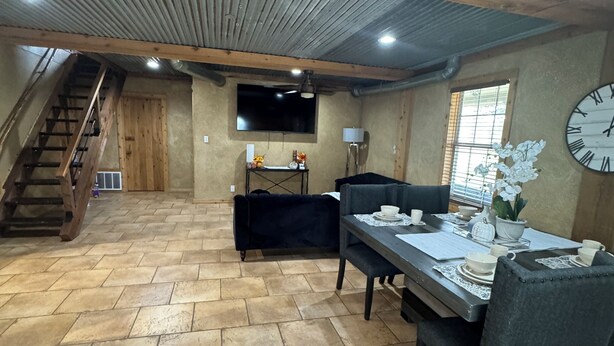Un vaste espace de salle à manger avec une table en bois dressée pour quatre personnes, un canapé noir et un escalier menant à un niveau supérieur.