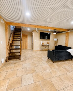 A spacious room with a wooden staircase, a black sofa, a television, and a ceiling fan.
